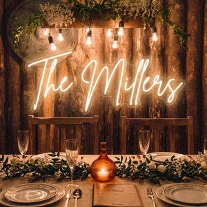 Puede incluir: Una mesa de recepci&oacute;n de boda con un letrero de ne&oacute;n que dice "The Millers". La mesa est&aacute; puesta con platos, cubiertos y vasos. Una vela en una botella de vidrio decorativa se encuentra en el centro. El fondo es una pared de madera.