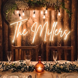May include: A warmly lit wedding reception table setting. The scene features a glowing neon sign that reads "The Millers" above a table set with plates, silverware, and champagne flutes. A decorative bottle with a candle inside sits in the center.