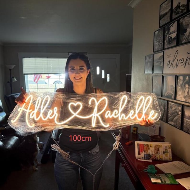 Puede incluir: Un letrero de ne&oacute;n con los nombres "Adler & Rachelle" y un coraz&oacute;n, iluminado en blanco c&aacute;lido. El letrero mide aproximadamente 100 cm de largo, ideal para decoraci&oacute;n interior.