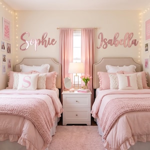 May include: Two twin beds with beige tufted headboards, pink bedding, and chunky knit blankets. Above each bed, large rose gold script wall signs read Sophie and Isabella. A white nightstand sits between the beds with a lamp and mirror.