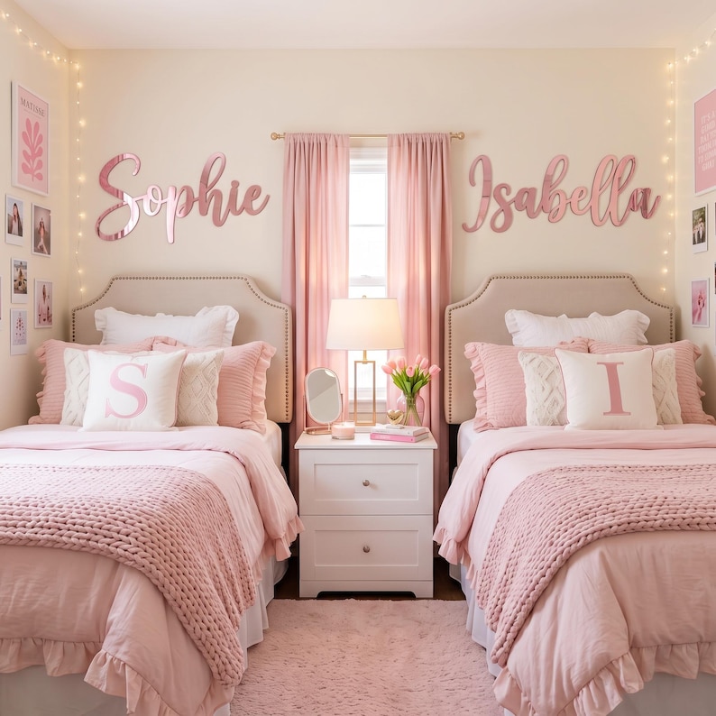 May include: Two twin beds with beige tufted headboards, pink bedding, and chunky knit blankets. Above each bed, large rose gold script wall signs read Sophie and Isabella. A white nightstand sits between the beds with a lamp and mirror.