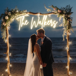 Puede incluir: Una pareja bes&aacute;ndose bajo un arco de madera decorado con flores y luces de cadena. El arco presenta un letrero de ne&oacute;n que dice "The Walkers". La escena est&aacute; ambientada en una playa al anochecer, con el oc&eacute;ano de fondo.