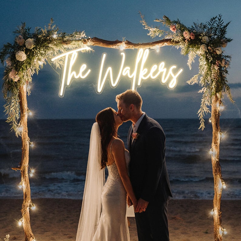 Puede incluir: Una pareja bes&aacute;ndose bajo un arco de madera decorado con flores y luces de cadena. El arco presenta un letrero de ne&oacute;n que dice "The Walkers". La escena est&aacute; ambientada en una playa al anochecer, con el oc&eacute;ano de fondo.