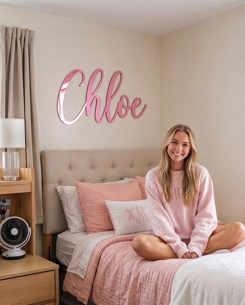May include: A large, cursive wall sign made of reflective pink material that spells out the name Chloe. The letters are connected in a single piece and mounted directly onto a plain, light-colored wall above a bed with pink bedding.