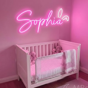 Puede incluir: Un letrero de ne&oacute;n rosa brillante que muestra el nombre Sophia en una fuente cursiva, montado en una pared rosa sobre una cuna blanca. La cuna contiene varios peluches y una manta tejida blanca y rosa sobre la barandilla lateral.