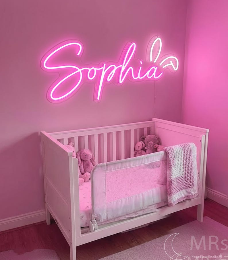 Puede incluir: Un letrero de ne&oacute;n rosa brillante que muestra el nombre Sophia en una fuente cursiva, montado en una pared rosa sobre una cuna blanca. La cuna contiene varios peluches y una manta tejida blanca y rosa sobre la barandilla lateral.
