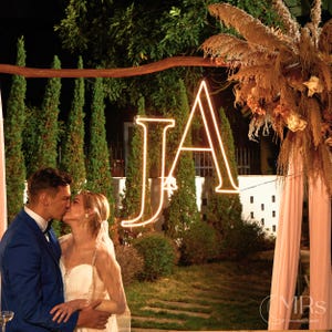 May include: A glowing neon sign featuring the letters JA in a serif font, suspended from a wooden archway decorated with dried pampas grass and light pink fabric, positioned outdoors at night with tall green cypress trees in the background.