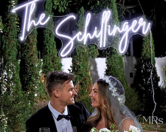 Puede incluir: Una recepción de boda con un letrero de neón blanco que dice "The Schiflinger" colgado sobre una pareja sentada en una mesa con flores blancas. La novia lleva un vestido blanco y velo, y el novio lleva un traje negro y corbata de moño.