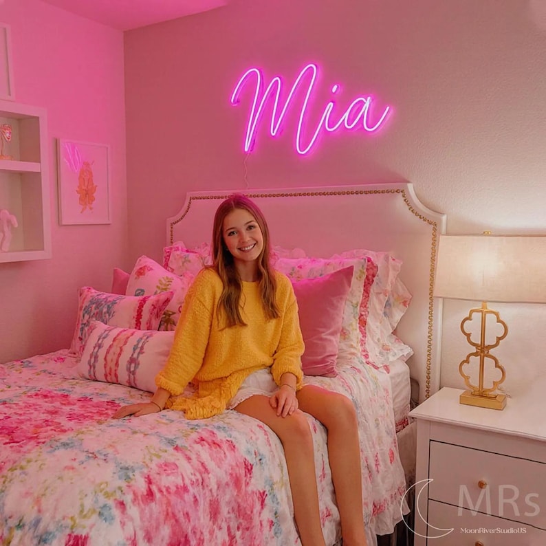 Puede incluir: Un letrero de ne&oacute;n rosa brillante montado en una pared que muestra el nombre Mia en una fuente cursiva. El letrero emite un brillo vibrante, iluminando el espacio sobre una cama con cabecera blanca, ropa de cama floral y una l&aacute;mpara de mesa dorada.