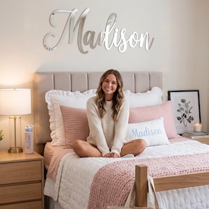 May include: A silver mirror-finish wall sign featuring the name Madison in a cursive script font. The sign is mounted on a wall above a bed. The name Madison is also printed in blue on a white rectangular pillow and on a light-colored water bottle.