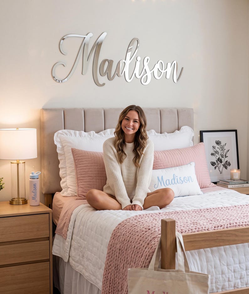 May include: A silver mirror-finish wall sign featuring the name Madison in a cursive script font. The sign is mounted on a wall above a bed. The name Madison is also printed in blue on a white rectangular pillow and on a light-colored water bottle.