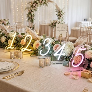 Puede incluir: Números de mesa de neón del 1 al 5, en amarillo, blanco y rosa, rodeados de un arreglo floral de rosas y vegetación. La mesa está preparada para una boda o evento.