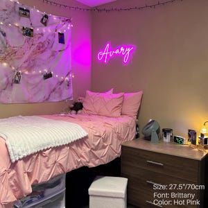 Puede incluir: Un letrero de ne&oacute;n rosa intenso que muestra el nombre Avary en una fuente cursiva Brittany, de 70 cm de ancho. El letrero est&aacute; montado en una pared sobre una cama con ropa de cama rosa, una manta blanca texturizada y un tapiz de pared de m&aacute;rmol.