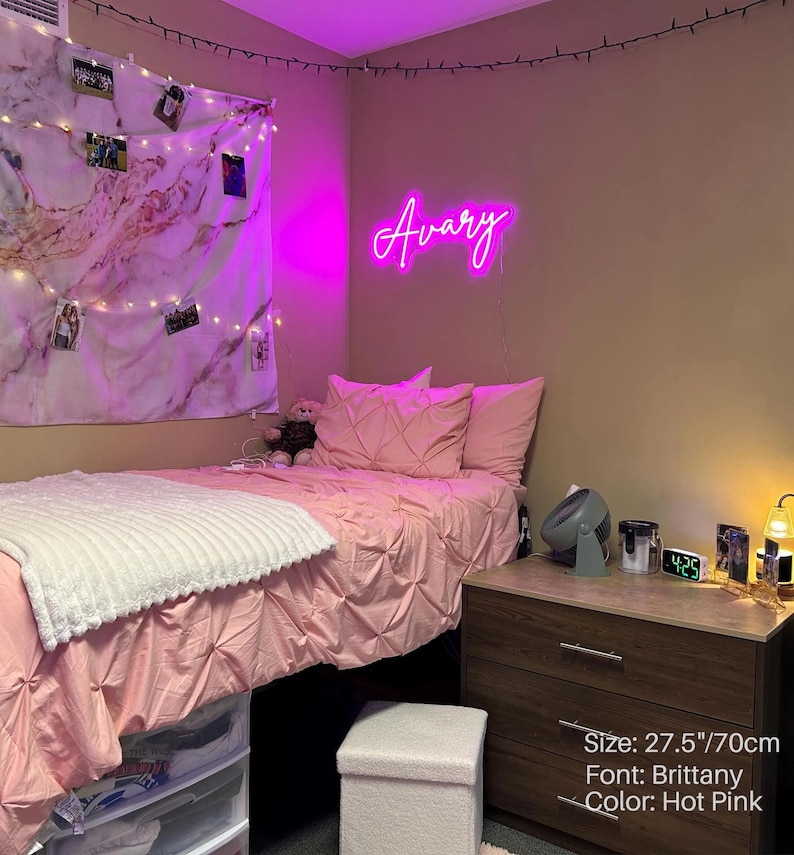Puede incluir: Un letrero de ne&oacute;n rosa intenso que muestra el nombre Avary en una fuente cursiva Brittany, de 70 cm de ancho. El letrero est&aacute; montado en una pared sobre una cama con ropa de cama rosa, una manta blanca texturizada y un tapiz de pared de m&aacute;rmol.