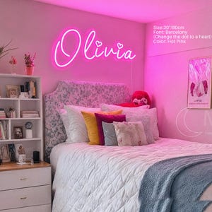 Puede incluir: Un letrero de ne&oacute;n rosa intenso que muestra el nombre Olivia en una fuente cursiva con puntos en forma de coraz&oacute;n sobre las letras i. El letrero mide 80 cm de ancho y est&aacute; montado en una pared sobre una cama con cabecero estampado y cojines.