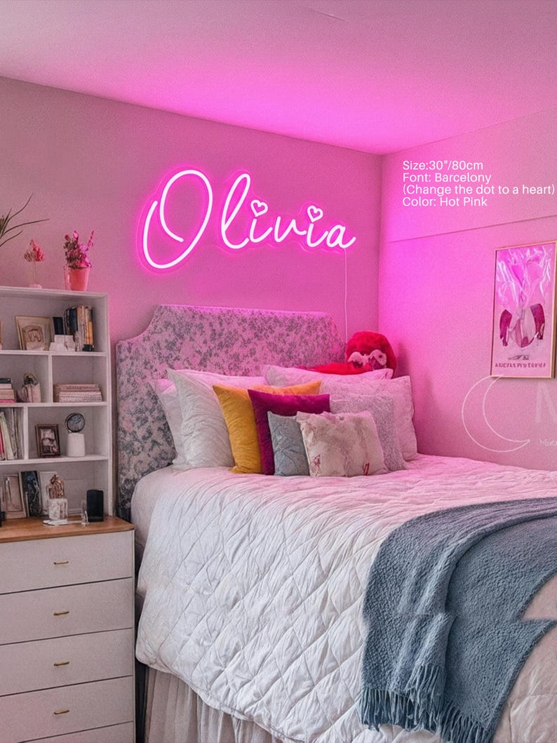 Puede incluir: Un letrero de ne&oacute;n rosa intenso que muestra el nombre Olivia en una fuente cursiva con puntos en forma de coraz&oacute;n sobre las letras i. El letrero mide 80 cm de ancho y est&aacute; montado en una pared sobre una cama con cabecero estampado y cojines.