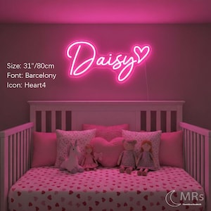 Puede incluir: Un letrero de ne&oacute;n rosa brillante que muestra el nombre Daisy en una fuente cursiva con un icono de coraz&oacute;n al final. El letrero mide 80 cm de ancho. El texto est&aacute; iluminado contra una pared oscura sobre una cuna con almohadas y mu&ntilde;ecas rosas.