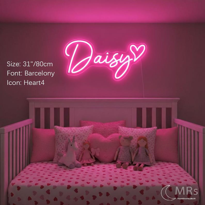 Puede incluir: Un letrero de ne&oacute;n rosa brillante que muestra el nombre Daisy en una fuente cursiva con un icono de coraz&oacute;n al final. El letrero mide 80 cm de ancho. El texto est&aacute; iluminado contra una pared oscura sobre una cuna con almohadas y mu&ntilde;ecas rosas.