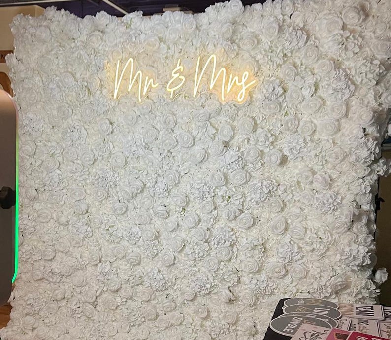 Puede incluir: Un tel&oacute;n de fondo de pared floral blanco con el letrero de ne&oacute;n "Mr & Mrs" en fuente cursiva. La pared est&aacute; densamente cubierta de rosas blancas, creando una superficie texturizada. Adecuado para bodas y eventos especiales.