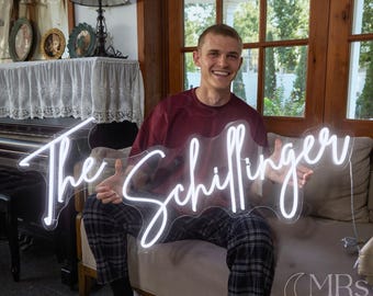 Könnte beinhalten: Ein weißes Neon-Schild mit dem Namen "The Schillinger" in Schreibschrift. Das Schild wird von einer Person gehalten. Das Schild befindet sich auf einer transparenten Rückseite mit weißem Rand. Das Schild ist beleuchtet und bereit zum Aufhängen.