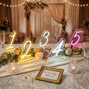Puede incluir: Decoración de mesa de boda con números iluminados del 1 al 5, rodeados de rosas y vegetación. Incluye copas de champán, velas y un letrero enmarcado que dice "Our Love Story Tables 1 to 5."