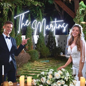 Puede incluir: Escena de boda con un letrero de ne&oacute;n que dice "The Martins". Una pareja est&aacute; de pie cerca de una mesa decorada con flores y velas. El hombre lleva un traje negro y pajarita, y la mujer un vestido de novia blanco y velo.