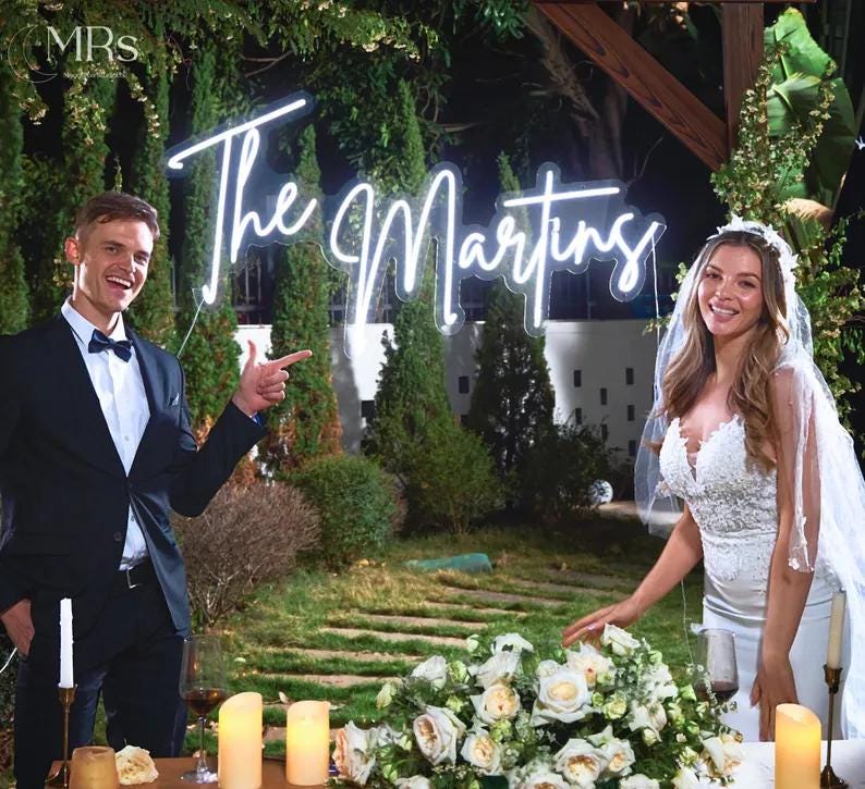 Puede incluir: Escena de boda con un letrero de ne&oacute;n que dice "The Martins". Una pareja est&aacute; de pie cerca de una mesa decorada con flores y velas. El hombre lleva un traje negro y pajarita, y la mujer un vestido de novia blanco y velo.
