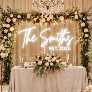 Puede incluir: Una mesa de recepci&oacute;n de bodas est&aacute; decorada con un mantel beige claro, adornada con arreglos florales en crema y burdeos. Un letrero de ne&oacute;n muestra "The Smiths EST. 2025". La escena est&aacute; enmarcada por arcos florales e iluminada por velas y una l&aacute;mpara de ara&ntilde;a.