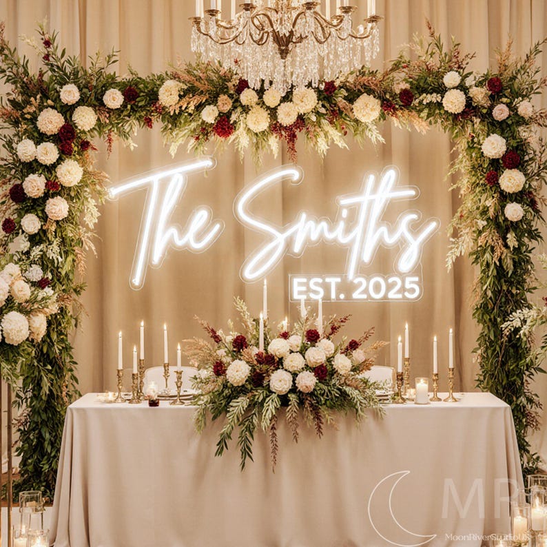 Puede incluir: Una mesa de recepci&oacute;n de bodas est&aacute; decorada con un mantel beige claro, adornada con arreglos florales en crema y burdeos. Un letrero de ne&oacute;n muestra "The Smiths EST. 2025". La escena est&aacute; enmarcada por arcos florales e iluminada por velas y una l&aacute;mpara de ara&ntilde;a.