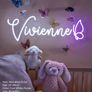 Puede incluir: Un letrero de ne&oacute;n en color blanco fr&iacute;o y morado que muestra el nombre Vivienne en fuente Monokind Script con un icono de mariposa, midiendo 60 cm de ancho. El letrero est&aacute; montado en una pared decorada con varias pegatinas de mariposas.
