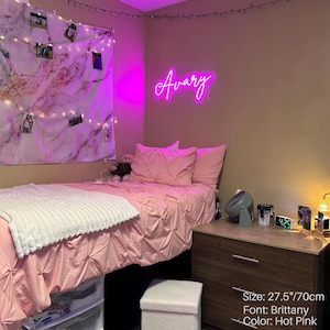 Puede incluir: Un letrero de ne&oacute;n rosa intenso que muestra el nombre Avary en una fuente cursiva Brittany, de 70 cm de ancho. El letrero est&aacute; montado en una pared sobre una cama con ropa de cama rosa, una manta blanca texturizada y un tapiz de pared de m&aacute;rmol.