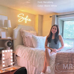 Puede incluir: Un letrero de ne&oacute;n blanco c&aacute;lido que muestra el nombre Sofia en fuente Barcelony, con un ancho de 60 cm. El letrero est&aacute; montado en una pared sobre una cama. El texto en la imagen incluye Size: 24"/60cm, Font: Barcelony, Color: Warm White.