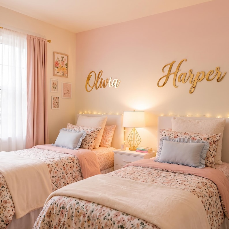 May include: Two twin beds with floral patterned bedding, solid light pink blankets, and light blue ruffled accent pillows. Above the beds, gold script wall signs read Olivia and Harper. A gold geometric lamp sits on a white nightstand.