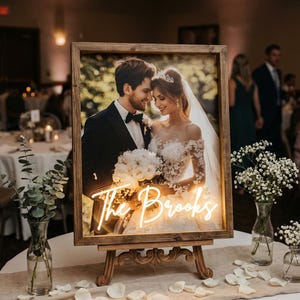 Puede incluir: Una foto de boda enmarcada con un letrero de ne&oacute;n que dice "The Brooks". La foto muestra a una pareja sonriendo. El marco es de madera y se exhibe en un caballete de madera. La mesa est&aacute; decorada con flores y p&eacute;talos.
