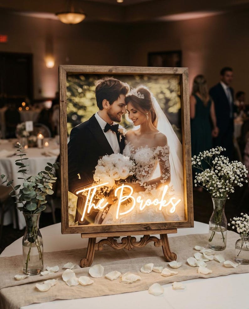 Puede incluir: Una foto de boda enmarcada con un letrero de ne&oacute;n que dice "The Brooks". La foto muestra a una pareja sonriendo. El marco es de madera y se exhibe en un caballete de madera. La mesa est&aacute; decorada con flores y p&eacute;talos.