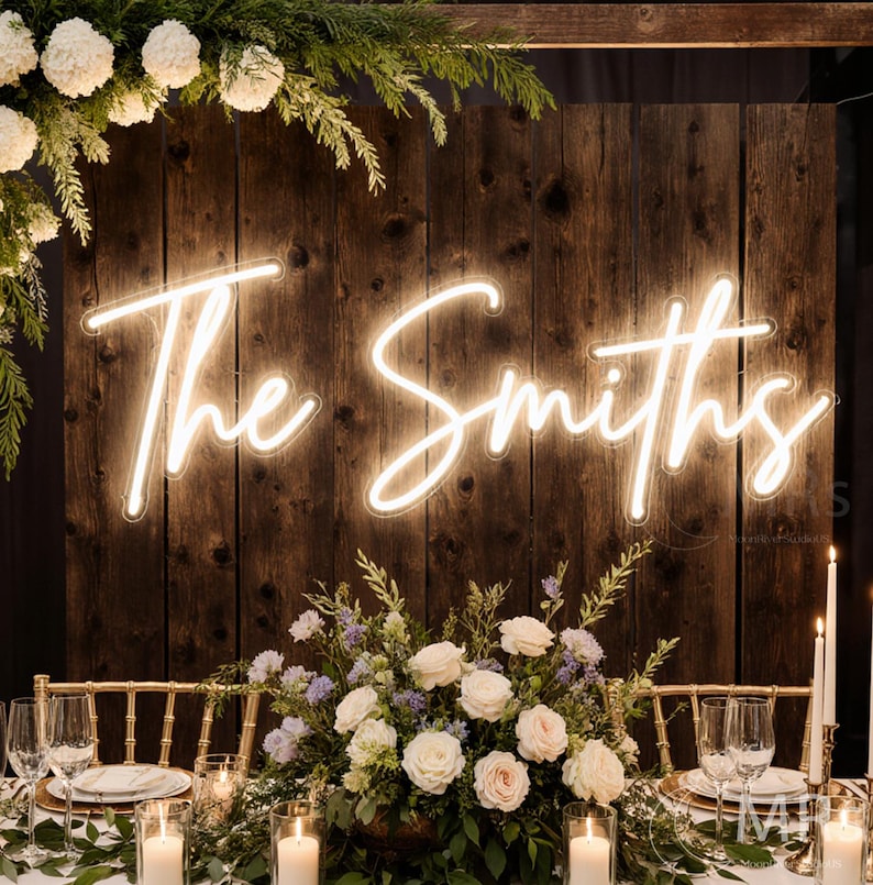 Puede incluir: Una mesa de recepci&oacute;n de bodas con un tel&oacute;n de fondo de madera con un letrero de ne&oacute;n que dice "The Smiths". La mesa est&aacute; decorada con flores blancas, velas y vegetaci&oacute;n. Sillas doradas y cubiertos completan el look elegante.