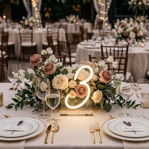 May include: A table setting with a floral centerpiece and a neon number 3. The centerpiece includes roses and greenery. The table is set with plates, glasses, and gold cutlery. Other tables are visible in the background.