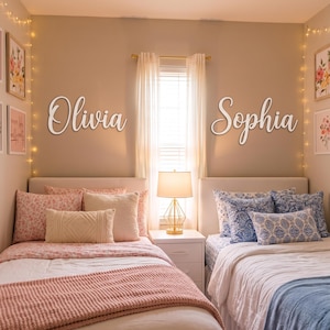 May include: Two white script wall signs featuring the names Olivia and Sophia mounted on a beige wall above two twin beds. The left bed has pink floral bedding and a pink throw, while the right bed has blue patterned bedding and a blue throw.