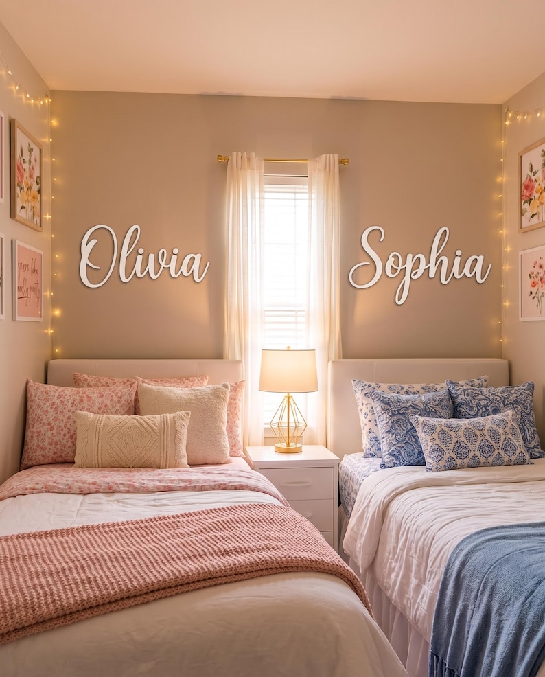 May include: Two white script wall signs featuring the names Olivia and Sophia mounted on a beige wall above two twin beds. The left bed has pink floral bedding and a pink throw, while the right bed has blue patterned bedding and a blue throw.