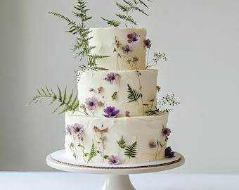 May include: A three-tiered white cake decorated with purple flowers and green fern leaves. The cake sits on a white pedestal cake stand. The cake has a textured buttercream frosting.