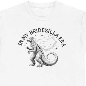 Puede incluir: Camiseta blanca con una ilustración en blanco y negro de una criatura parecida a Godzilla que lleva un velo de novia. El texto "In My Bridezilla Era" está escrito en un círculo alrededor de la criatura.