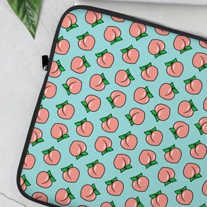 Puede incluir: Una funda para portátil con un patrón repetido de melocotones rosas con hojas verdes sobre un fondo azul claro. La funda tiene un borde negro y un cierre de cremallera. El portátil es parcialmente visible.