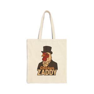 Puede incluir: Bolso tote de color crema con un dibujo de un pollo de dibujos animados con sombrero de copa, gafas y pajarita. El texto "Chicken Zaddy" está impreso en naranja y marrón. El bolso tiene asas largas y un diseño sencillo.