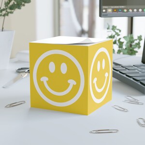 Può includere: Un blocco note giallo a forma di cubo con una faccia sorridente bianca su ogni lato. Il blocco note è su una superficie bianca.