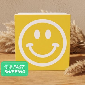 May include: A square yellow wooden block featuring a white outline of a classic smiley face with two oval eyes and a curved mouth centered on the front surface. The block has a smooth finish and is displayed in a bright, clear orientation.