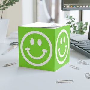 Puede incluir: Un dispensador de notas adhesivas de color verde en forma de cubo con una carita sonriente blanca en cada lado. El dispensador está sobre una superficie blanca.
