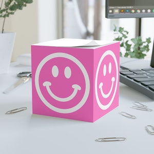 Könnte beinhalten: Rosa, würfelförmiger Klebezettelspender mit einem weißen Smiley auf jeder Seite.
