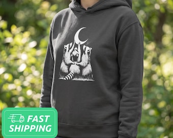 May include: A charcoal gray hooded sweatshirt featuring a white graphic print of three raccoons standing on their hind legs and reaching up toward a crescent moon. The long-sleeved pullover includes a hood with drawstrings and ribbed cuffs.