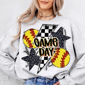 Może przedstawiać: Szara bluza z nadrukiem graficznym przedstawiającym dwa wzory piłek do softballa w kolorze ż&oacute;łtym i czerwonym, wz&oacute;r w biało-czarną kratę, czarną gwiazdę z nadrukiem lamparta i tekst "GAME DAY".