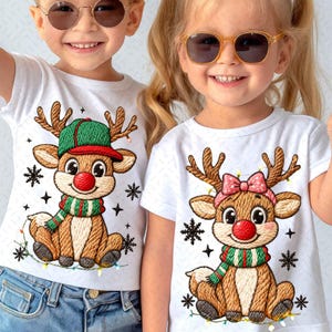 Op de afbeelding: Twee witte T-shirts met geborduurde rendierontwerpen. Eén rendier draagt een groene en rode hoed en sjaal, de andere een roze strik en groene sjaal. Beide hebben rode neuzen en sneeuwvlokken.